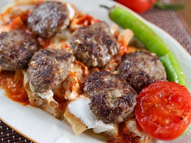 Een bord Balaban-kebab: gehaktballetjes op brood met tomatensaus en yoghurt, met gegrilde tomaat en groene peper